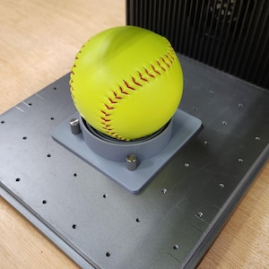 Könnte beinhalten: Ein gelber Softball mit roten Nähten liegt auf einer grauen Plattform mit einem grauen Ring darum.