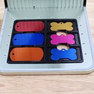 May include: A metal tag making machine with a tray containing six blank metal tags. The tags are in different shapes and colours, including red, blue, orange, gold, pink, and light blue.