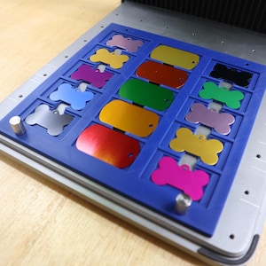 Op de afbeelding: Een blauwe bak met een raster van 12 botvormige metalen labels in verschillende kleuren, waaronder rood, oranje, geel, groen, blauw, paars en zilver. De labels zijn in een rasterpatroon op de bak gerangschikt.