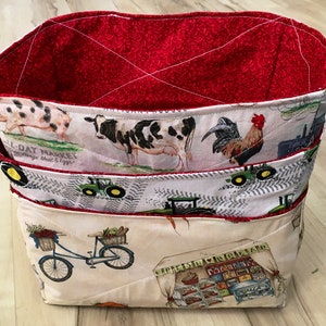 May include: Three fabric storage baskets with a farm theme. The top basket is red, the middle basket has a cow, pig, and rooster, and the bottom basket has a bicycle and a farm stand.