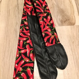 May include: A black and red fabric strap with a chili pepper pattern. The strap has two black leather ends with adjustable buckles.