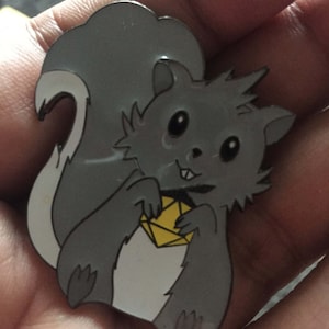 Puede incluir: Un pin de esmalte gris de una ardilla de dibujos animados sosteniendo un diamante amarillo. La ardilla tiene la barriga blanca y la cola tupida.
