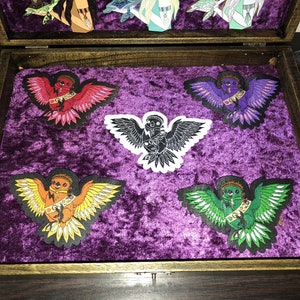 Peut inclure: Cinq patchs thermocollants en forme d'oiseau dans une boîte en bois avec un revêtement en velours violet. Les patchs présentent des oiseaux de différentes couleurs avec les ailes déployées et une couronne sur la tête.