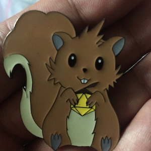 Puede incluir: Un pin de ardilla de dibujos animados marrón y blanco con un diamante amarillo en sus patas. La ardilla tiene una cola marrón y ojos negros.