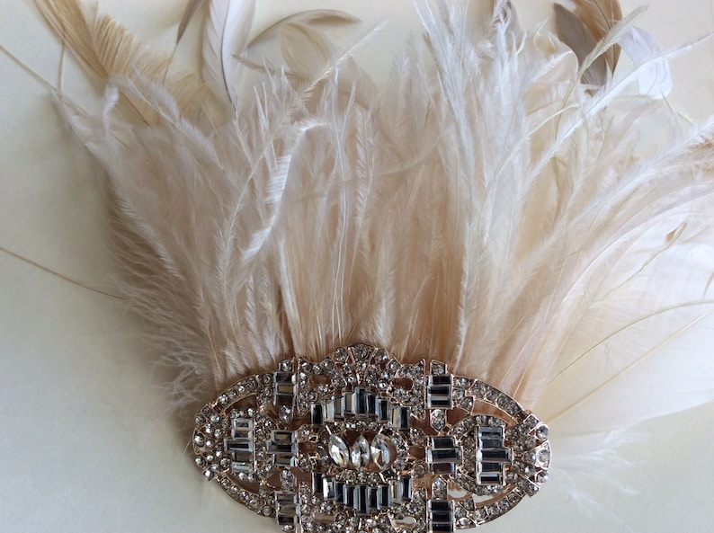 K&ouml;nnte beinhalten: Ein Kopfschmuck im Vintage-Stil mit einem F&auml;cher aus cremefarbenen Federn. Die Federn sind an einem ovalen, goldfarbenen Metallst&uuml;ck befestigt, das mit klaren Strasssteinen und Kristallen besetzt ist. F&uuml;r besondere Anl&auml;sse.