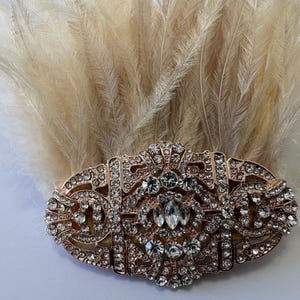 K&ouml;nnte beinhalten: Haarspange im Art-Deco-Stil in Ros&eacute;gold, verziert mit klaren Strasssteinen und einem B&uuml;schel beiger Federn. Die Spange hat ein ovales Design mit aufwendigen Details und einem zentralen, gr&ouml;&szlig;eren Strassstein.