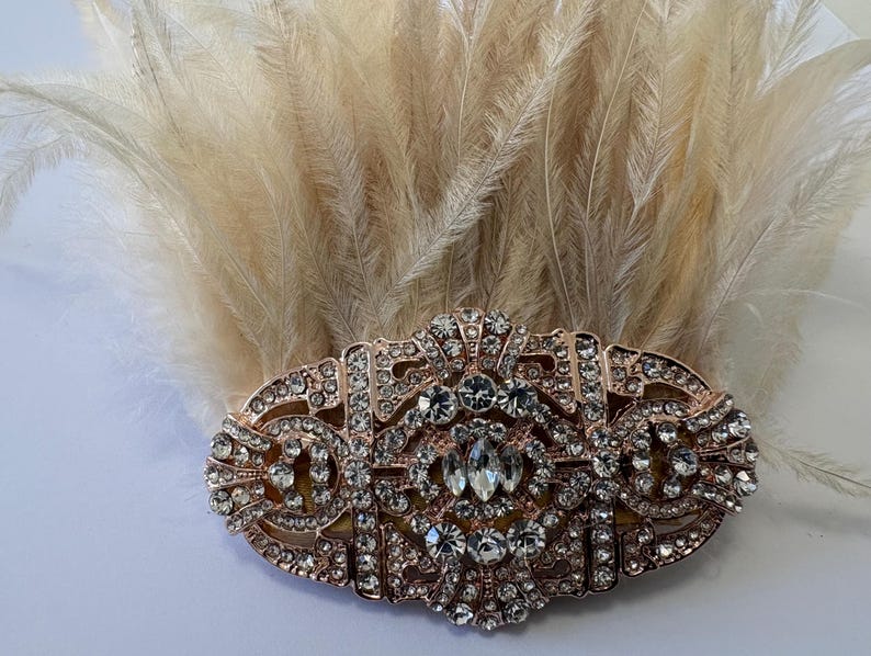 K&ouml;nnte beinhalten: Haarspange im Art-Deco-Stil in Ros&eacute;gold, verziert mit klaren Strasssteinen und einem B&uuml;schel beiger Federn. Die Spange hat ein ovales Design mit aufwendigen Details und einem zentralen, gr&ouml;&szlig;eren Strassstein.