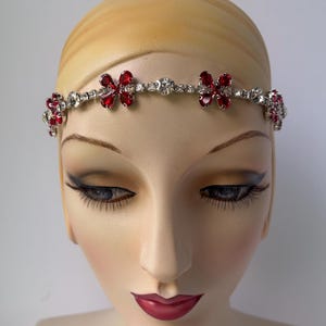 May include: A silver-toned headband adorned with red and clear crystal flower accents. The floral design is set against a row of clear crystals. The headband is displayed on a mannequin head with painted features.