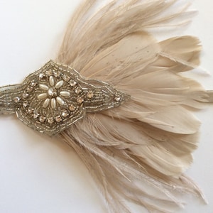 May include: A beige feather headband featuring a central decorative applique. The applique is embellished with clear rhinestones, silver beads, and small white pearls arranged in a floral pattern on a light-colored fabric base.