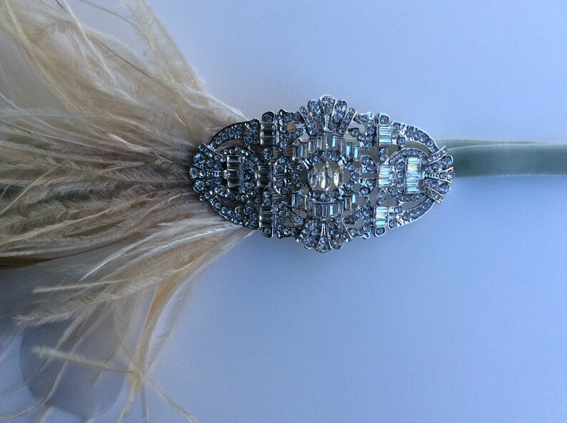 K&ouml;nnte beinhalten: Ein Kopfschmuck im Art-Deco-Stil mit einem silberfarbenen, verzierten Mittelst&uuml;ck, das mit klaren Strasssteinen besetzt ist. Weiche, cremefarbene Federn fallen von der Seite herab, befestigt an einem hellgr&uuml;nen Samtband. Ein glamour&ouml;ses Accessoire.