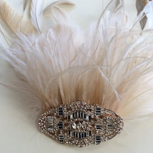 K&ouml;nnte beinhalten: Ein Kopfschmuck im Vintage-Stil mit einem F&auml;cher aus cremefarbenen und beigefarbenen Federn. Die Federn sind &uuml;ber einer verzierten, silberfarbenen Brosche angeordnet, die mit funkelnden Strasssteinen besetzt ist. Die Brosche hat ein rechteckiges und ovales Design.