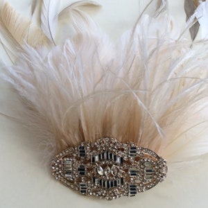 K&ouml;nnte beinhalten: Ein Kopfschmuck im Vintage-Stil mit einem F&auml;cher aus cremefarbenen und beigefarbenen Federn. Die Federn sind &uuml;ber einer ovalen, silberfarbenen Brosche mit funkelnden Strasssteinen angeordnet. Geeignet f&uuml;r besondere Anl&auml;sse.
