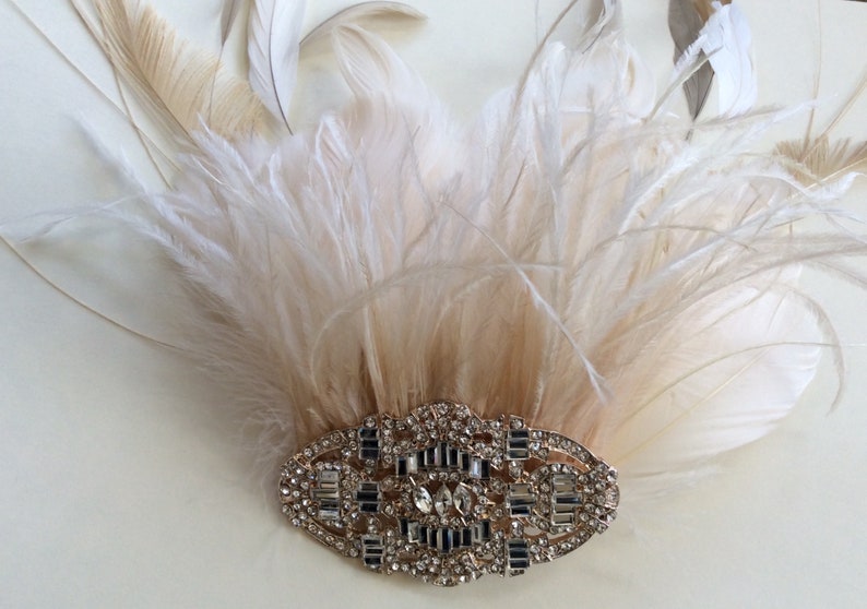 K&ouml;nnte beinhalten: Ein Kopfschmuck im Vintage-Stil mit einem F&auml;cher aus cremefarbenen und beigefarbenen Federn. Die Federn sind &uuml;ber einer ovalen, silberfarbenen Brosche mit funkelnden Strasssteinen angeordnet. Geeignet f&uuml;r besondere Anl&auml;sse.