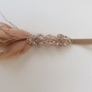 May include: A delicate headband featuring a decorative accent with sparkling rhinestones and small beads in a diamond shape. The headband also has soft, light brown feathers attached to the side. The band itself is a light brown color.