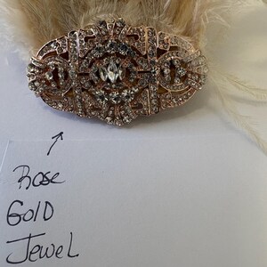 K&ouml;nnte beinhalten: Haarschmuck in Ros&eacute;gold-Optik mit klaren Strasssteinen. Das ovale St&uuml;ck hat ein verziertes Design und ist vor einem Hintergrund aus hellen Federn angeordnet. Die Worte "Rose Gold Jewel" sind auf einem St&uuml;ck Papier geschrieben.