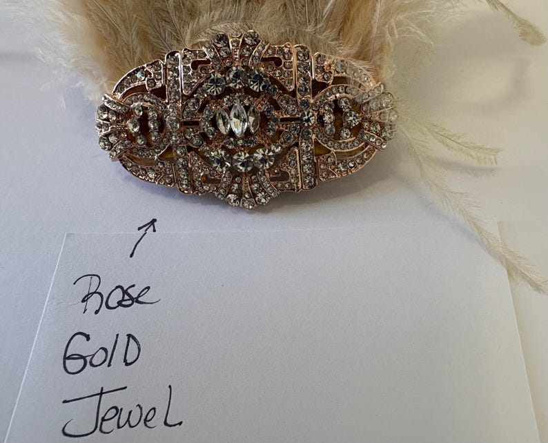 K&ouml;nnte beinhalten: Haarschmuck in Ros&eacute;gold-Optik mit klaren Strasssteinen. Das ovale St&uuml;ck hat ein verziertes Design und ist vor einem Hintergrund aus hellen Federn angeordnet. Die Worte "Rose Gold Jewel" sind auf einem St&uuml;ck Papier geschrieben.
