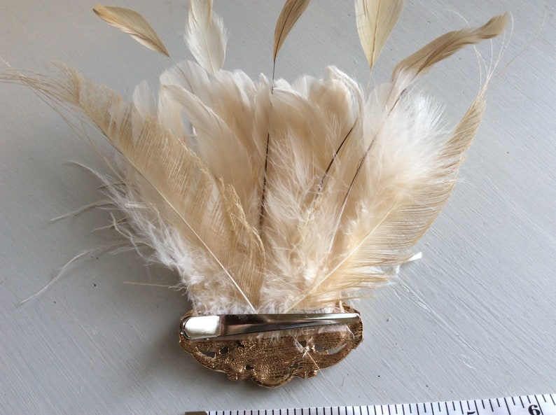 K&ouml;nnte beinhalten: Dekorative Haarspange mit einem F&auml;cher aus cremefarbenen Federn und einem goldfarbenen Metallclip. Die Federn sind halbkreisf&ouml;rmig angeordnet, wobei einige Federn nach oben ragen. Die Spange ist etwa 10 cm lang.