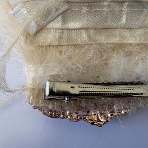 K&ouml;nnte beinhalten: Elfenbeinfarbene Haarspange mit Metallverschluss und dekorativen Verzierungen. Die Spange ist mit weichen, flauschigen Federn und einem Satinband verziert, wodurch ein zartes und elegantes Accessoire entsteht. Zum Befestigen der Haare.