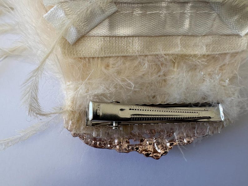 K&ouml;nnte beinhalten: Elfenbeinfarbene Haarspange mit Metallverschluss und dekorativen Verzierungen. Die Spange ist mit weichen, flauschigen Federn und einem Satinband verziert, wodurch ein zartes und elegantes Accessoire entsteht. Zum Befestigen der Haare.