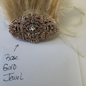 K&ouml;nnte beinhalten: Ros&eacute;goldenes Schmuckst&uuml;ck mit klaren Strasssteinen, vor einem Hintergrund aus hellen Federn. Das Schmuckst&uuml;ck hat ein verziertes, symmetrisches Design. Der Text auf dem Papier lautet "Rose Gold Jewel".