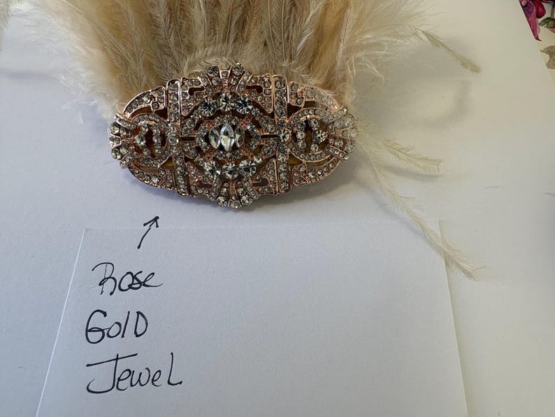 K&ouml;nnte beinhalten: Ros&eacute;goldenes Schmuckst&uuml;ck mit klaren Strasssteinen, vor einem Hintergrund aus hellen Federn. Das Schmuckst&uuml;ck hat ein verziertes, symmetrisches Design. Der Text auf dem Papier lautet "Rose Gold Jewel".