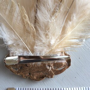 K&ouml;nnte beinhalten: Dekorative Haarspange mit einem F&auml;cher aus cremefarbenen Federn. Die Federn sind an einer goldfarbenen Metallspange mit einem dekorativen Design befestigt. Die Spange ist etwa 7.6 cm lang.