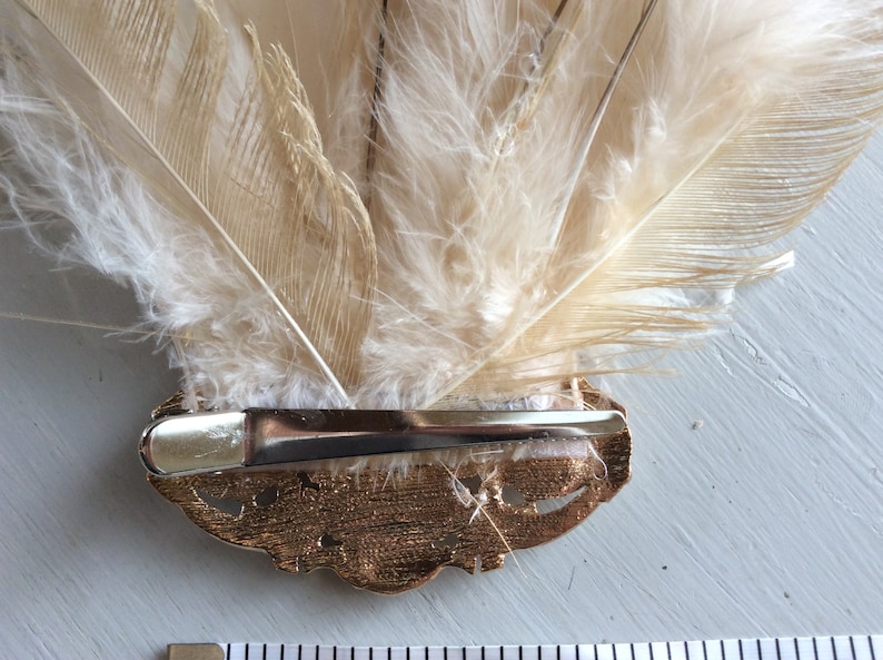 K&ouml;nnte beinhalten: Dekorative Haarspange mit einem F&auml;cher aus cremefarbenen Federn. Die Federn sind an einer goldfarbenen Metallspange mit einem dekorativen Design befestigt. Die Spange ist etwa 7.6 cm lang.