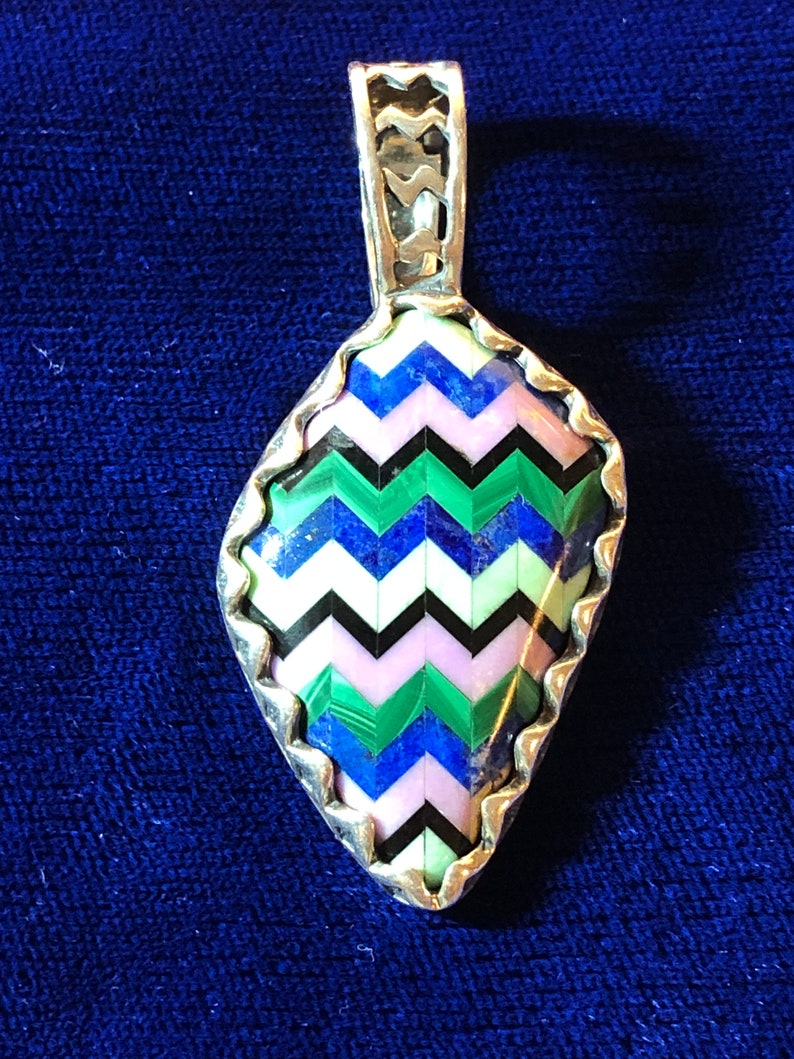 Puede incluir: Un colgante de plata con un dise&ntilde;o geom&eacute;trico que presenta una variedad de piedras de colores, incluyendo azul, verde, rosa y negro.