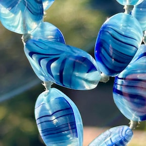 Pode incluir: Close-up de um colar com contas ovais azuis. As contas translúcidas apresentam riscas onduladas azuis escuras. O colar é enfiado num cordão de cor clara. As contas medem cerca de 2 cm de comprimento.