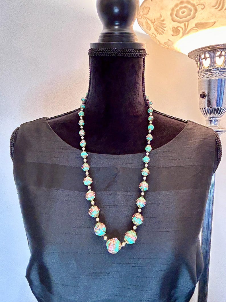 May include: A turquoise and pink beaded necklace with gold accents. The beads are round and have a floral design. The necklace is on a black mannequin.