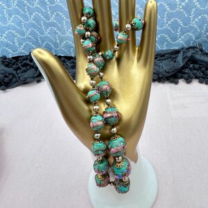May include: A long necklace made of turquoise blue glass beads with gold and pink accents. The beads are round and have a textured surface. The necklace is displayed on a gold hand-shaped stand.