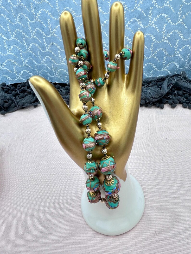 May include: A long necklace made of turquoise blue glass beads with gold and pink accents. The beads are round and have a textured surface. The necklace is displayed on a gold hand-shaped stand.