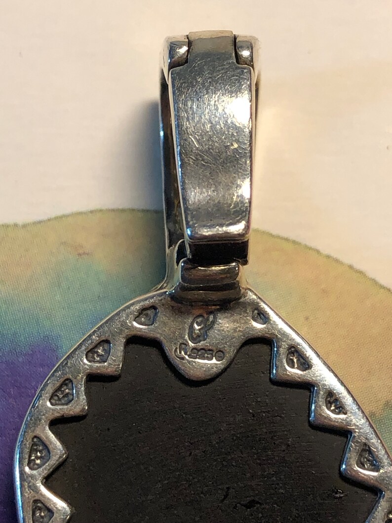Puede incluir: Primer plano de un colgante de plata con una piedra negra. El colgante tiene un dise&ntilde;o detallado alrededor del borde y un cierre de plata. La parte posterior del colgante est&aacute; estampada con las letras "CP" y el n&uacute;mero "9250".