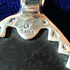 Puede incluir: Primer plano de un colgante de plata con una piedra negra. El colgante tiene un dise&ntilde;o detallado y est&aacute; marcado con las letras "GF" y "925".