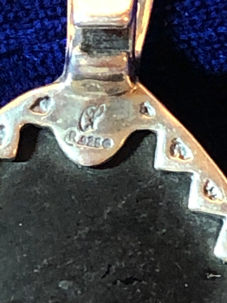 Puede incluir: Primer plano de un colgante de plata con una piedra negra. El colgante tiene un dise&ntilde;o detallado y est&aacute; marcado con las letras "GF" y "925".