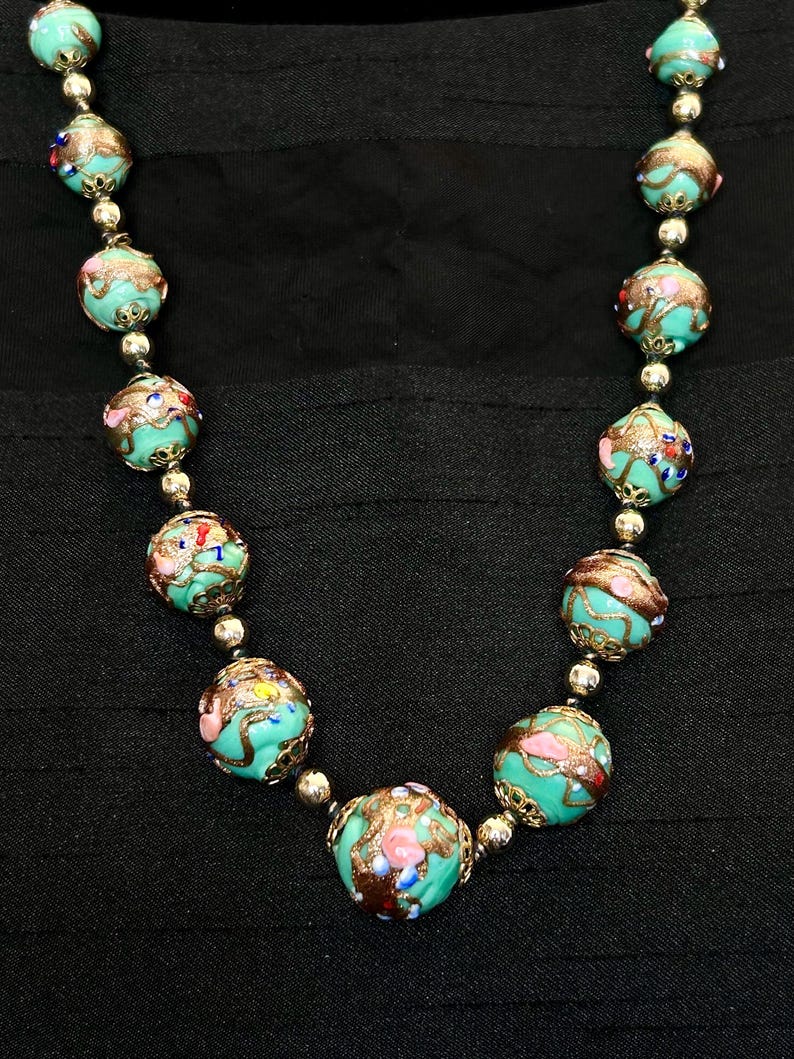 May include: A turquoise and gold necklace with round beads. The beads are decorated with intricate floral patterns and colorful accents.