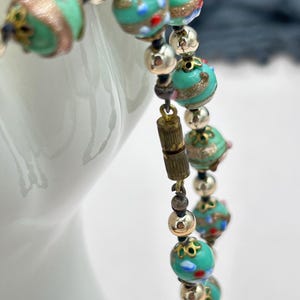 May include: A turquoise and gold beaded necklace with a silver clasp. The beads are made of glass and have a variety of patterns and colors.
