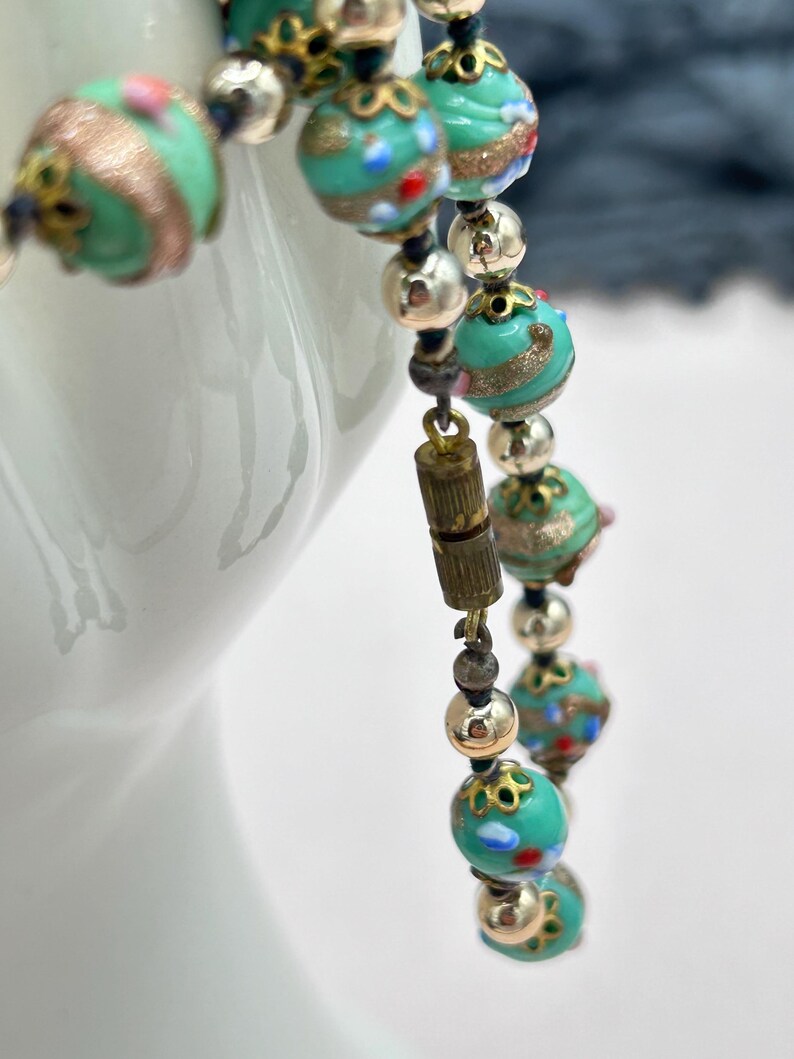 May include: A turquoise and gold beaded necklace with a silver clasp. The beads are made of glass and have a variety of patterns and colors.