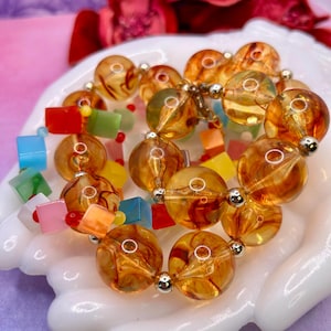 May include: A vintage necklace featuring large, amber-colored, marbled glass beads interspersed with smaller silver beads. The necklace also includes colorful square and rectangular beads in shades of red, blue, green, and yellow. The necklace is displayed on a white, hand-shaped dish.