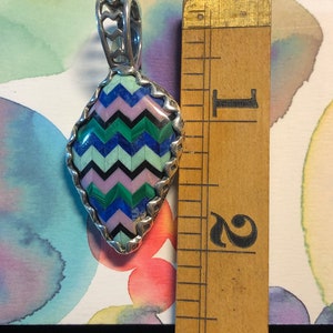 Puede incluir: Un colgante de plata con un dise&ntilde;o geom&eacute;trico hecho de piedras turquesa, rosa, verde y azul. El colgante mide aproximadamente 3,8 cm de alto y se encuentra sobre un fondo blanco con una regla para la escala.