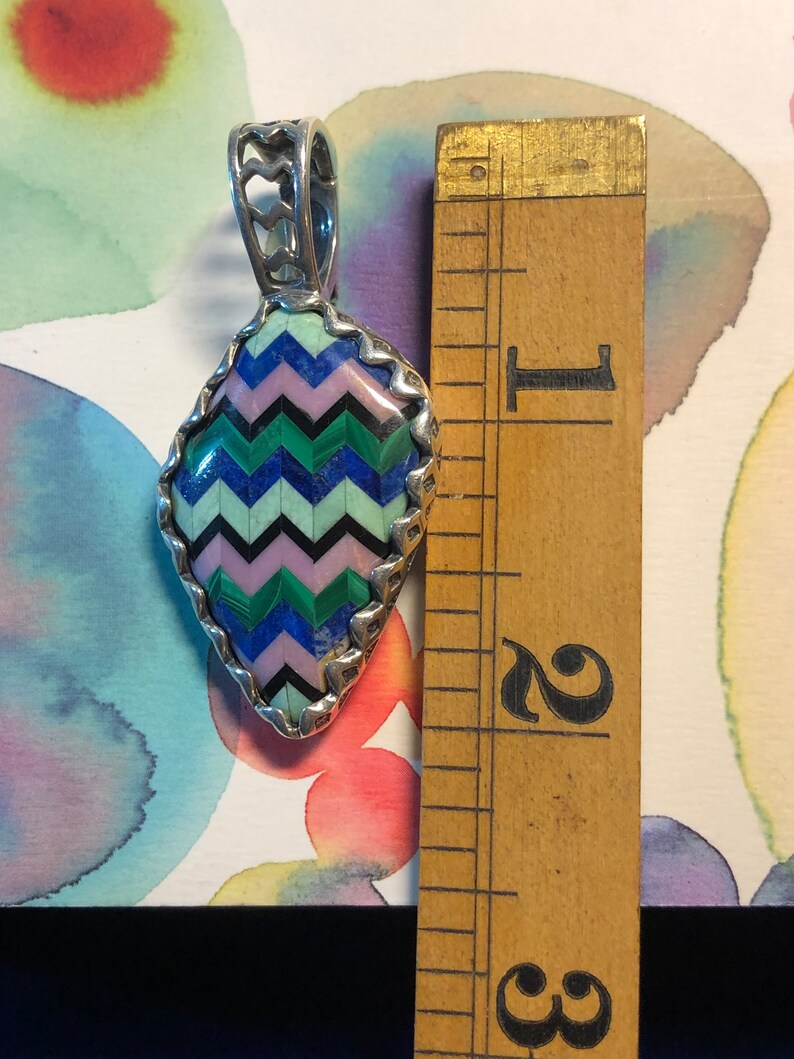Puede incluir: Un colgante de plata con un dise&ntilde;o geom&eacute;trico hecho de piedras turquesa, rosa, verde y azul. El colgante mide aproximadamente 3,8 cm de alto y se encuentra sobre un fondo blanco con una regla para la escala.
