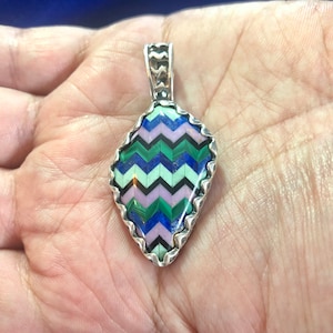 Puede incluir: Un colgante de plata con un patr&oacute;n de chevron hecho de varias piedras, incluyendo verde, azul, rosa y negro. El colgante tiene forma de l&aacute;grima y tiene un borde festoneado.