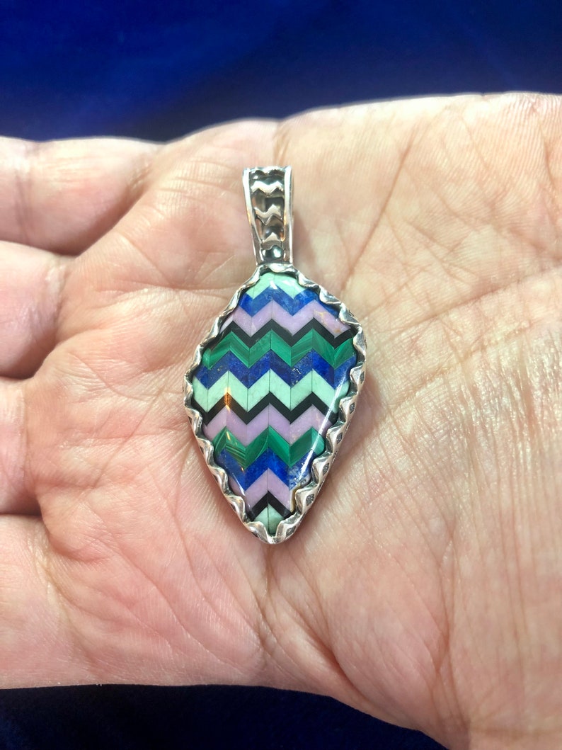 Puede incluir: Un colgante de plata con un patr&oacute;n de chevron hecho de varias piedras, incluyendo verde, azul, rosa y negro. El colgante tiene forma de l&aacute;grima y tiene un borde festoneado.