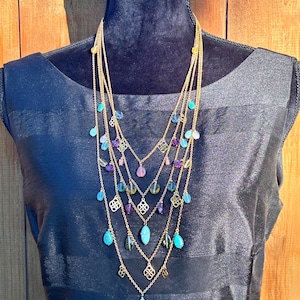 May include: A multi-strand gold-coloured necklace with turquoise, purple, and clear gemstone accents. The necklace features a layered design with decorative filigree elements and teardrop-shaped pendants. The necklace is displayed on a black dress form.