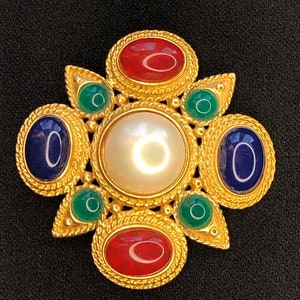 Op de afbeelding: Een gouden broche met een groot parel middenstuk omringd door kleinere rode, blauwe en groene stenen.