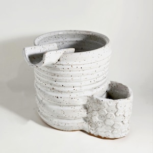 May include: White ceramic planter with a unique design featuring a spout and a honeycomb-shaped side. The planter has a speckled glaze and a rustic, handmade look.