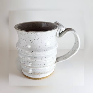 May include: White ceramic mug with a speckled glaze and a unique, twisted handle. The mug has a rounded body with a slightly flared rim.