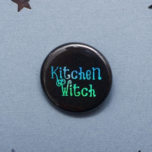 Puede incluir: Un botón negro con un texto degradado en verde y azul que dice "Kitchen Witch".