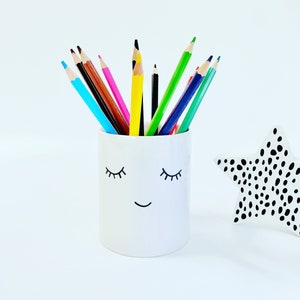 May include: A white ceramic pencil holder with a smiling face and eyelashes. The holder is filled with coloured pencils, including blue, green, yellow, orange, red, and brown.
