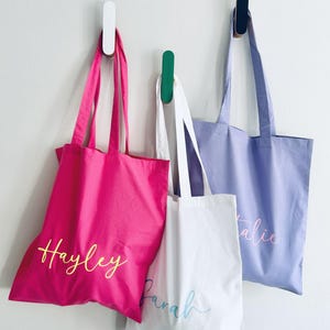 May include: Three tote bags hanging on hooks. The bag on the left is pink with the name "Hayley" written in yellow. The middle bag is white with the name "Sarah" written in blue. The bag on the right is light purple with the name "Talie" written in pink.